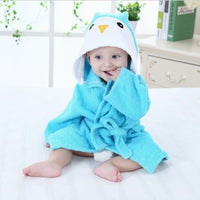 Baby in blauem Kapuzenhandtuch mit Pinguin-Design, sitzt auf weißem Bett, niedlich und kuschelig.
