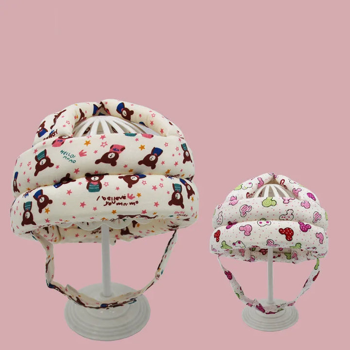 Zwei Babyhelme mit Bären- und Wolkenmuster, beige und rosa, auf Ständern vor rosa Hintergrund.