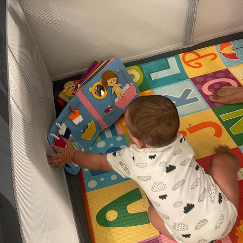 Kleinkind spielt mit buntem Stoffbuch auf Alphabet-Spielmatte im Laufstall.