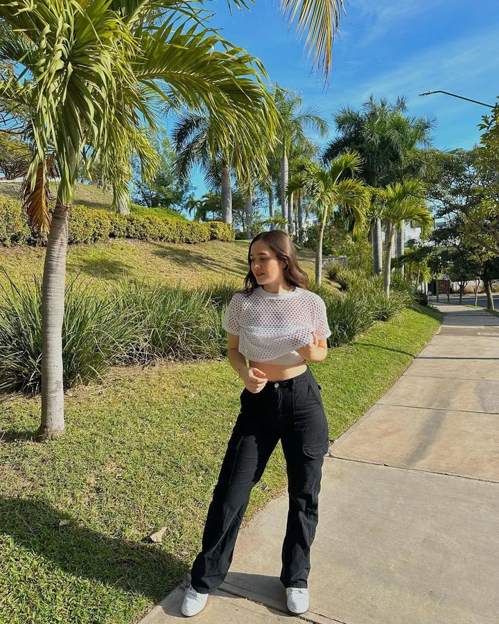 Frau in weißem Netzoberteil und schwarzer Hose spaziert im sonnigen Park mit Palmen.