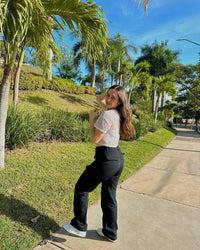 Junge Frau in weißem Spitzenoberteil und schwarzer Hose spaziert auf sonnigem Gehweg.