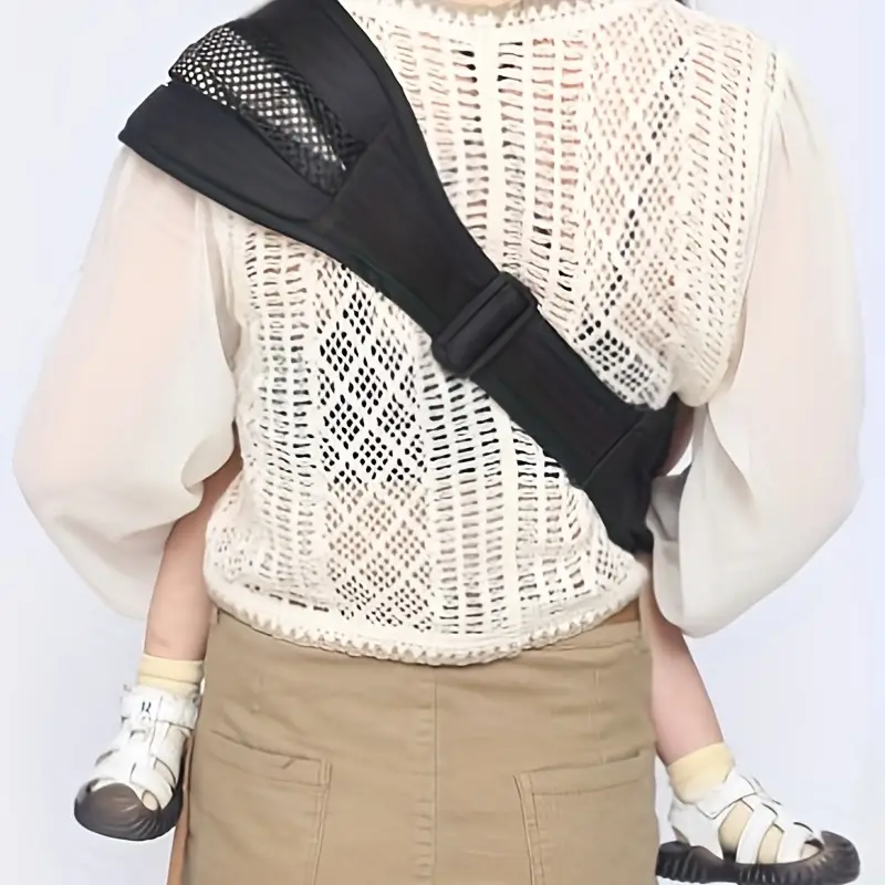 Person mit Babytrage, schwarzer Schultergurt, beige Kleidung, Baby in weißen Schuhen.