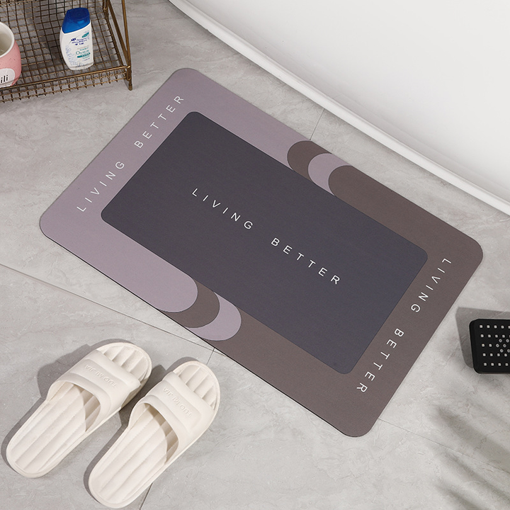 Graue Badematte mit "Living Better" Schriftzug auf Fliesenboden, weiße Hausschuhe daneben.