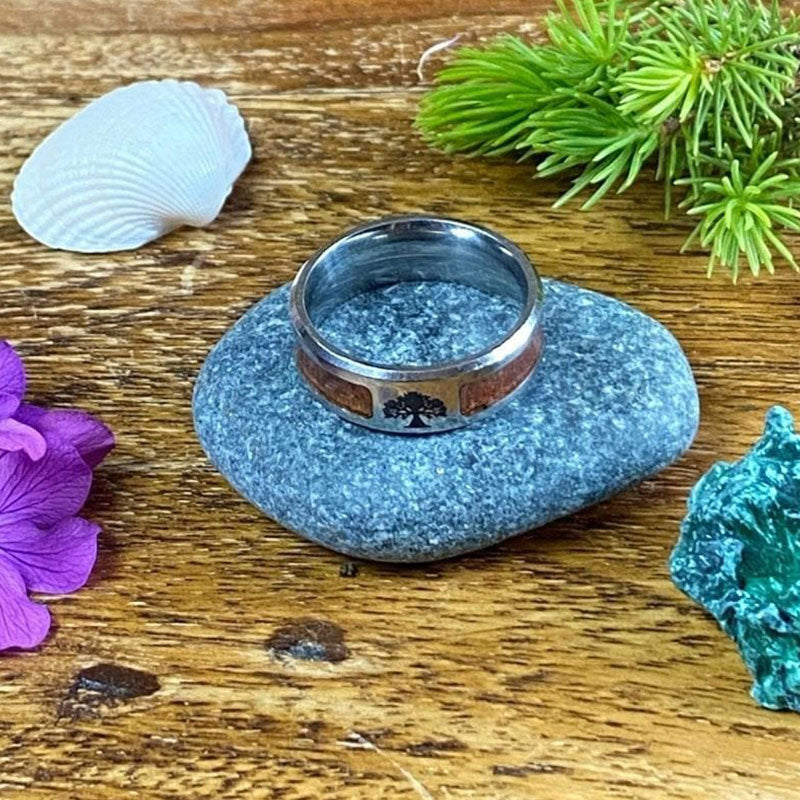 Silberring mit Holzintarsien auf Stein, umgeben von Muscheln und Blumen, rustikaler Hintergrund.