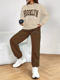 Frau in beigem Brooklyn-Sweatshirt und brauner Jogginghose, hält schwarzen Becher, weiße Sneaker.