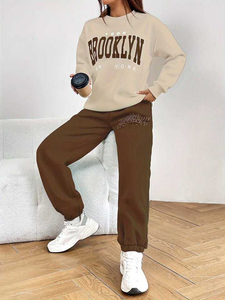 Frau in beigem Brooklyn-Sweatshirt und brauner Jogginghose, hält schwarzen Becher, weiße Sneaker.