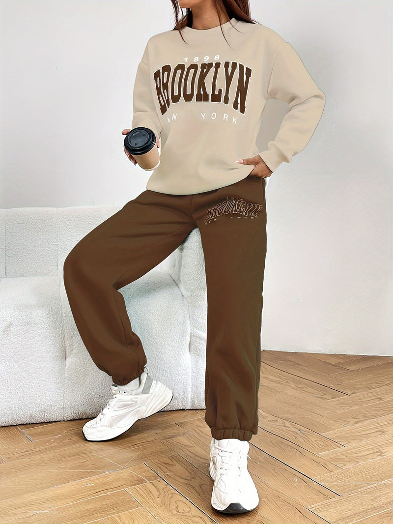 Frau in beigem Brooklyn-Sweatshirt und brauner Jogginghose, hält schwarzen Becher, weiße Sneaker.