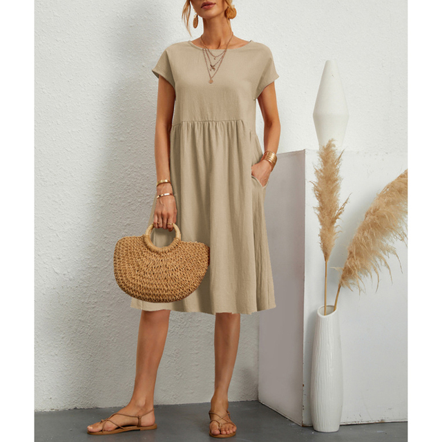 Frau in beige Baumwoll-Leinen-Kleid, Boho-Stil, mit Strohtasche und Sandalen, vor weißer Wand.