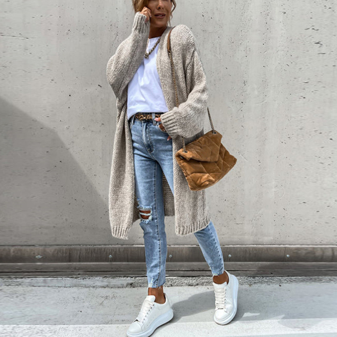 Frau in beigem Strickcardigan, blauer Jeans, weißen Sneakers, brauner Tasche vor Betonwand.