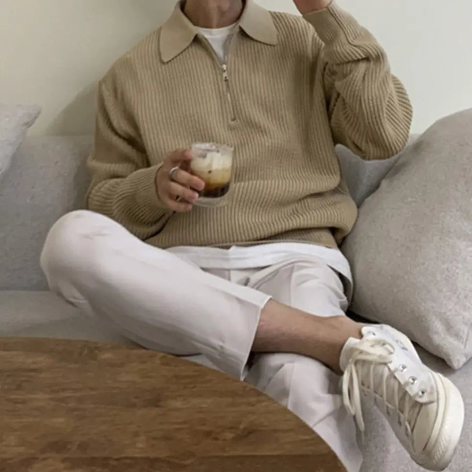 Mann in beigem Strickpullover mit Reißverschluss, hält Kaffee, sitzt auf Sofa, weiße Hose, Sneaker.