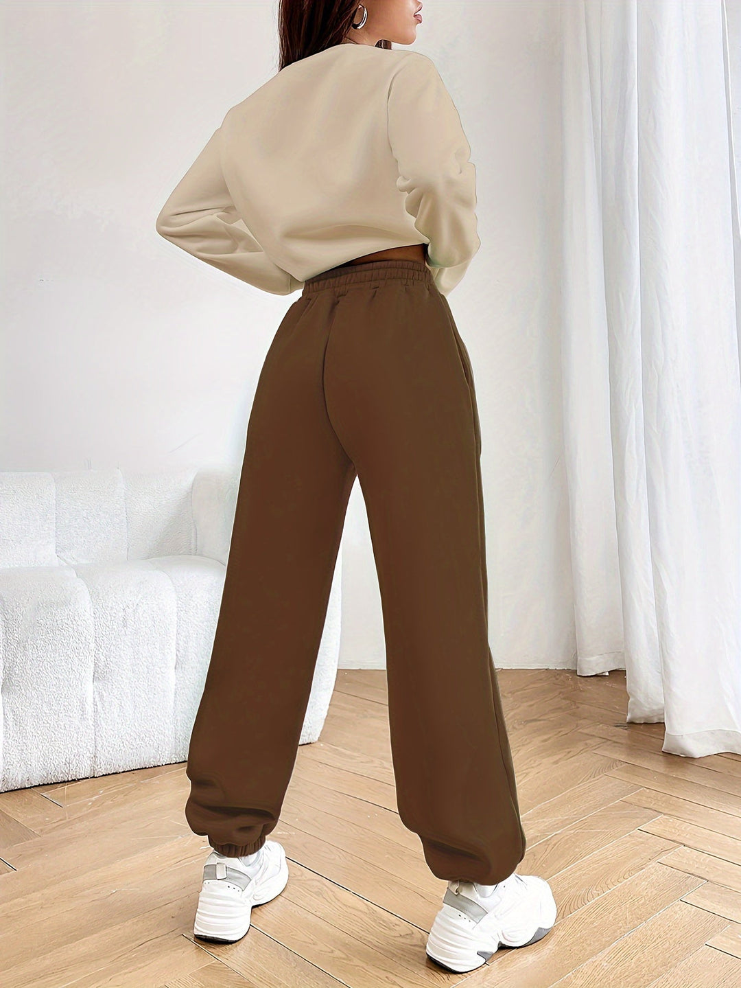 Frau in beiger Bluse und brauner Jogginghose, weiße Sneaker, modernes Wohnzimmer, Holzboden.