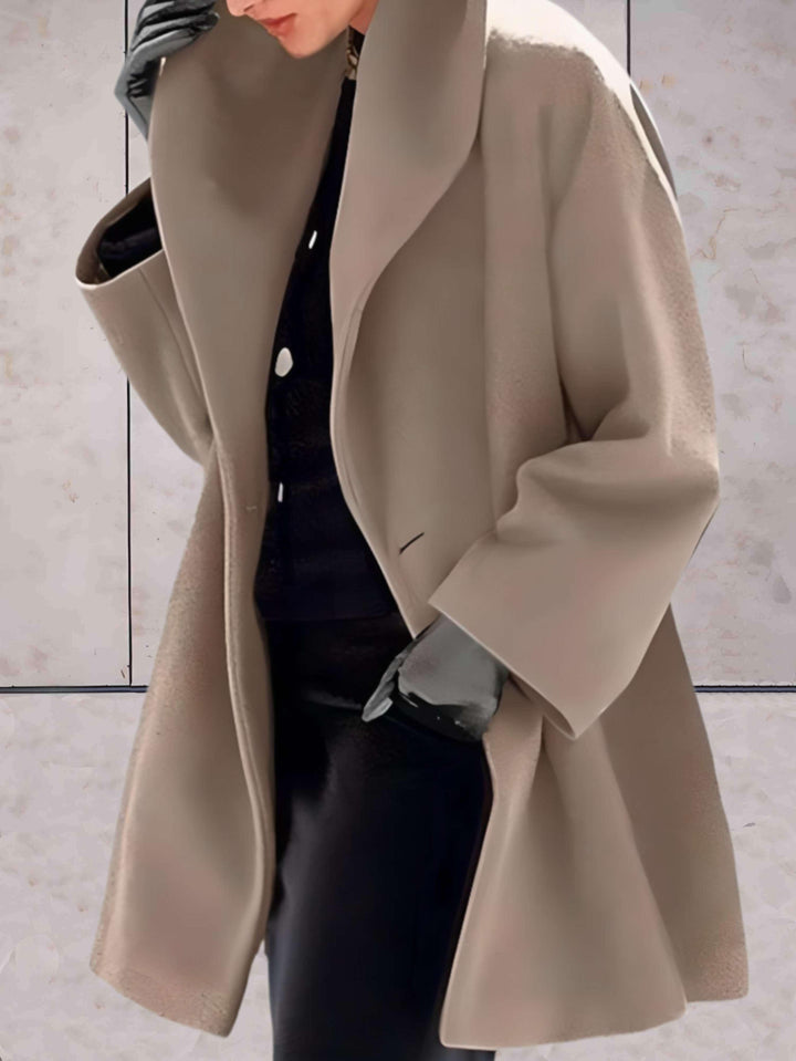 Frau in beigem Wollmantel, schwarzer Kleidung und Handschuhen vor Marmorhintergrund.