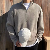 Mann in beigem Strickpullover mit Reißverschluss hält weiße Baseballkappe vor Holzwand.