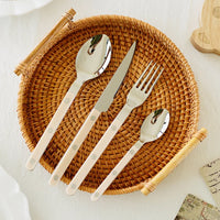 Besteckset aus Edelstahl mit cremefarbenen Griffen auf einem runden, geflochtenen Tablett.