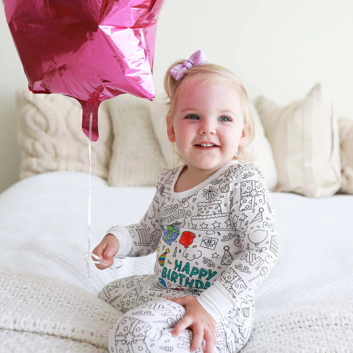 Kleinkind im weißen Pyjama mit "Happy Birthday"-Aufdruck hält rosa Sternballon auf Bett.