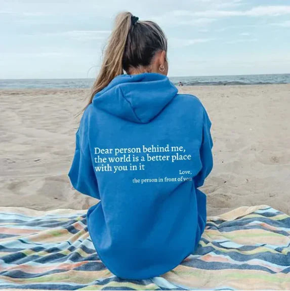 Frau am Strand in blauem Hoodie mit motivierendem Zitat, sitzt auf gestreifter Decke.