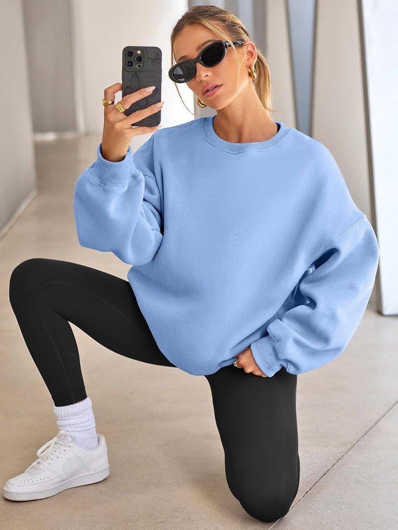 Frau in hellblauem Sweatshirt, schwarze Leggings, weiße Sneaker, Sonnenbrille, Smartphone.
