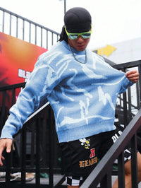 Mann in blauem Strickpullover mit abstraktem Muster, Sonnenbrille, schwarze Shorts, Streetwear-Look.