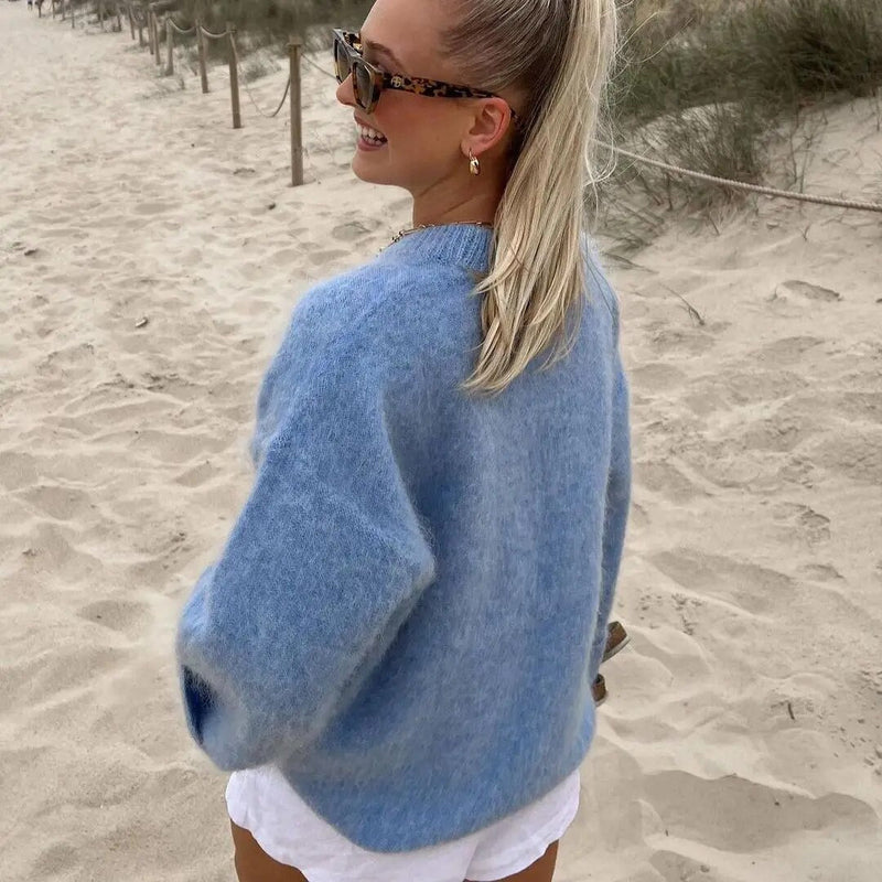 Frau am Strand in blauem Strickpullover, weiße Shorts, Sonnenbrille, blonder Pferdeschwanz.
