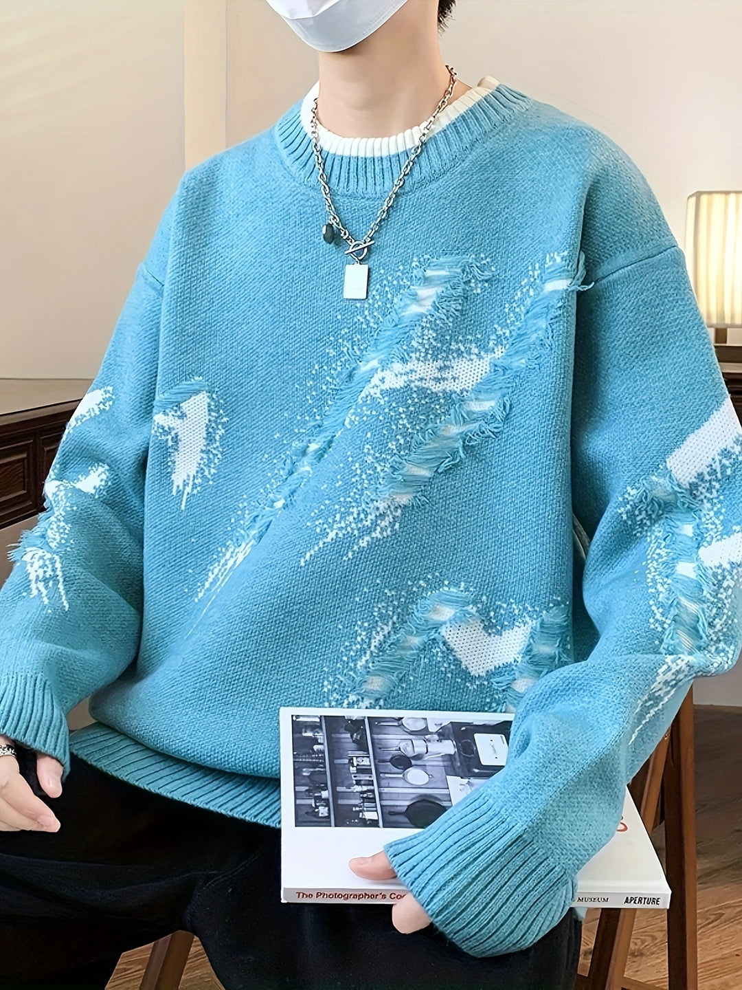 Person in blauem Strickpullover mit abstraktem Muster, hält ein Buch, modisch, lässiger Stil.