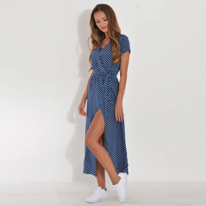 Frau im blauen gepunkteten Maxikleid mit Schlitz und weißen Sneakers vor weißem Hintergrund.