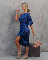 Frau in blauem Satin-Kleid, einseitig schulterfrei, mit beiger Handtasche und Sandalen.