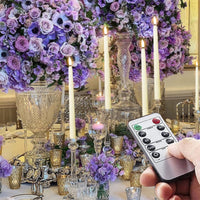 Hand hält Fernbedienung vor elegantem Tisch mit lila Blumen und Kerzenständern.