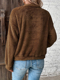 Frau in brauner Fleecejacke und blauer Jeans vor Holztür und weißer Ziegelwand.