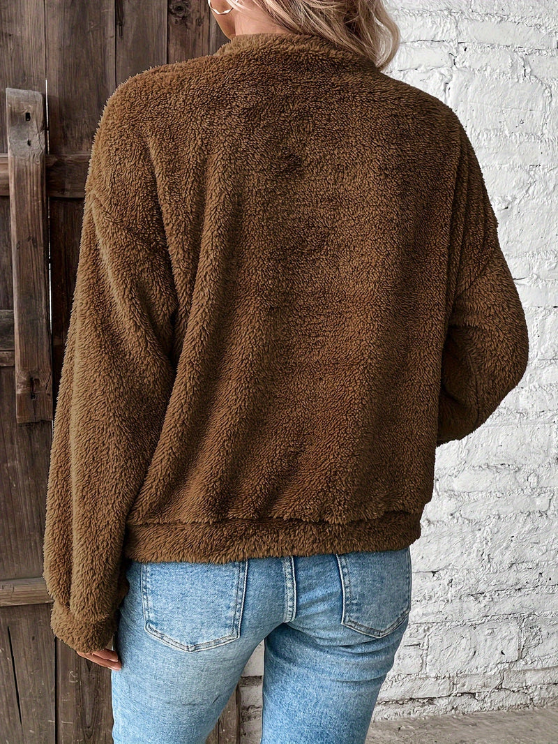 Frau in brauner Fleecejacke und blauer Jeans vor Holztür und weißer Ziegelwand.