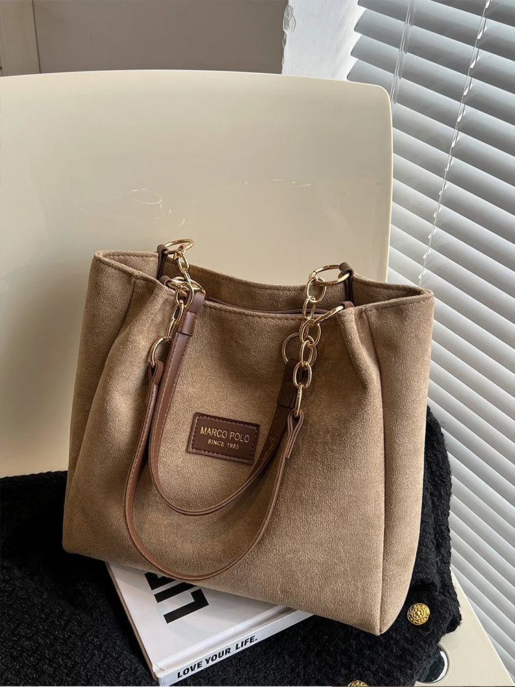 Beige Damenhandtasche aus Wildleder mit goldenen Kettenriemen auf weißem Stuhl.