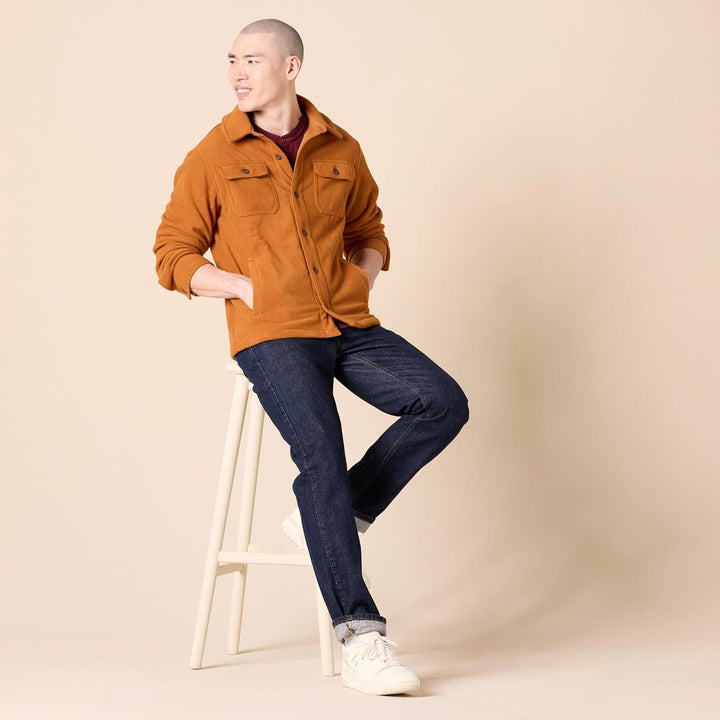 Mann in brauner Jacke und blauer Jeans sitzt auf Hocker, lächelt, beige Hintergrund.