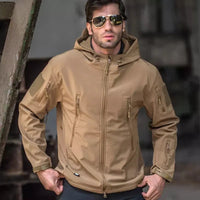 Mann in brauner Outdoor-Softshelljacke mit Kapuze und Reißverschlusstaschen, trägt Sonnenbrille.