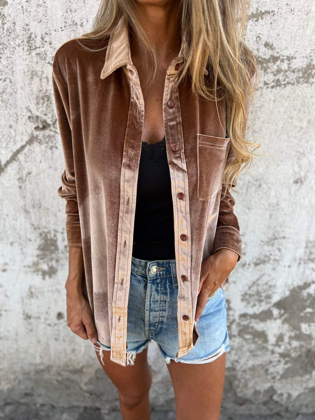 Frau in brauner Samtjacke, schwarzem Top und blauen Jeansshorts vor Betonwand.