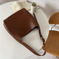 Braune Ledertasche auf weißem Tisch, neben Buch und Kaffeetasse, moderne Einrichtung.