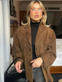 Frau in brauner Wildlederjacke, schwarzem Rollkragenpullover, mit schwarzer Tasche im Hotelzimmer.