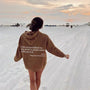 Frau am Strand in braunem Hoodie mit Spruch, Sonnenuntergang, Sand, Freizeitkleidung.