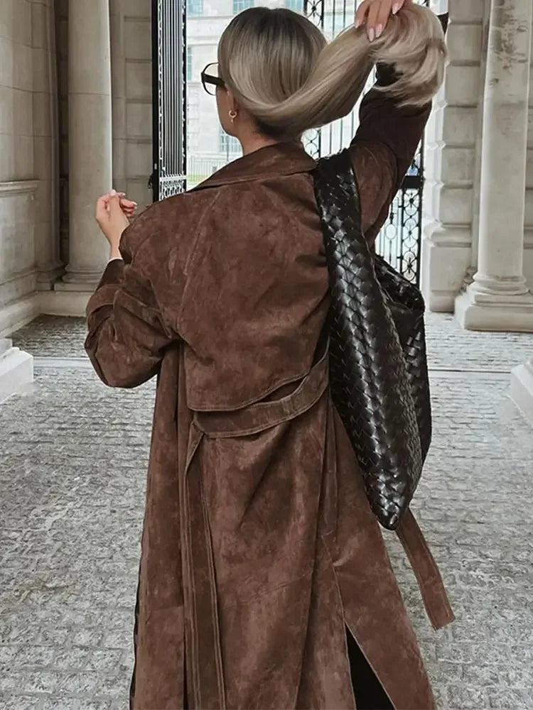 Frau mit braunem Wildledermantel und schwarzer geflochtener Tasche in urbaner Umgebung.