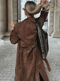 Frau mit braunem Wildledermantel und schwarzer geflochtener Tasche in urbaner Umgebung.