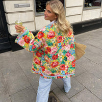 Frau in bunter Blumenjacke, gestreifte Hose, mit Getränk, auf Gehweg, Sommermode.