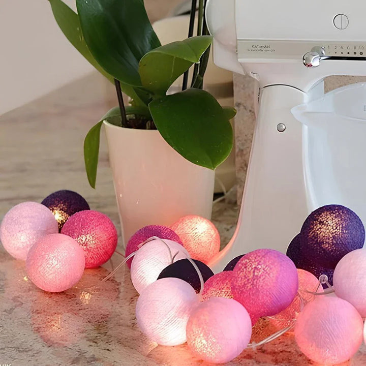 Bunte Lichterkette mit rosa und lila Kugeln auf Küchentheke neben weißem Mixer und Pflanze.