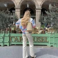 Frau mit Patchwork-Jacke und Strohtasche vor Café mit grüner Fassade und Olivenbäumen.
