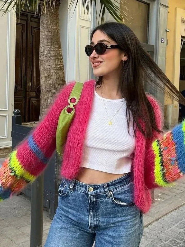 Frau in bunter Strickjacke, weißem Top, blauer Jeans, Sonnenbrille, grüne Tasche, Straße.