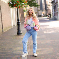 Frau in bunter Strickjacke, blauer Jeans, lila Tasche, weiße Schuhe, Stadtstraße, Blumenampel.