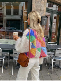 Frau mit buntem Strickpullover, weißer Hose, brauner Tasche, Kaffeebecher, draußen im Café.