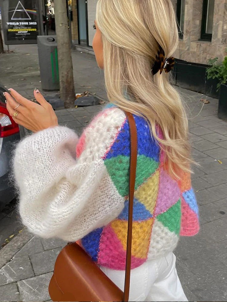 Frau mit buntem Strickpullover, weißer Hose, brauner Tasche, auf städtischer Straße.