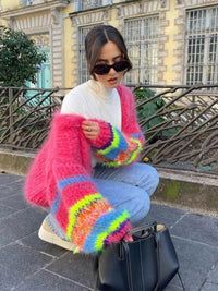 Frau mit pinkem, buntem Strickpullover, Sonnenbrille, Jeans, kniend neben schwarzer Tasche.