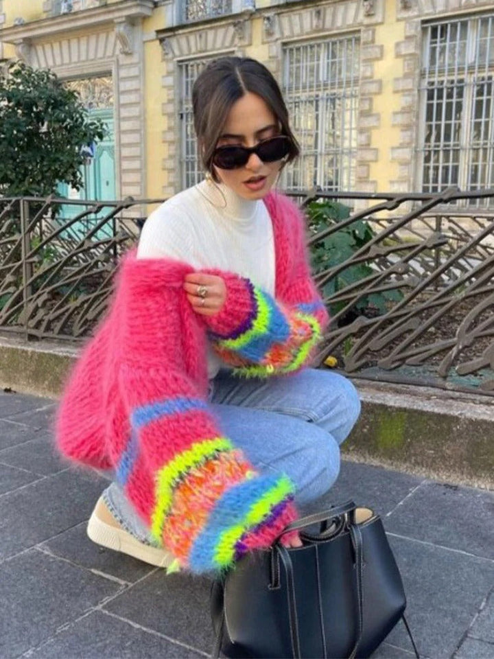 Frau mit pinkem, buntem Strickpullover, Sonnenbrille, Jeans, kniend neben schwarzer Tasche.