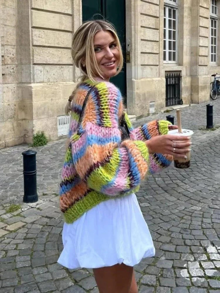 Frau in bunter Strickjacke, weißem Kleid, hält Bubble Tea, steht auf Kopfsteinpflasterstraße.