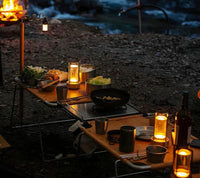 Camping-Tisch mit Beleuchtung, Geschirr und Essen am Flussufer bei Nacht, Outdoor-Abenteuer.