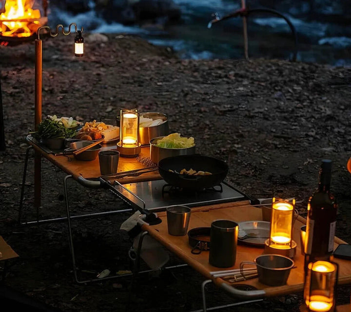 Camping-Tisch mit Beleuchtung, Geschirr und Essen am Flussufer bei Nacht, Outdoor-Abenteuer.